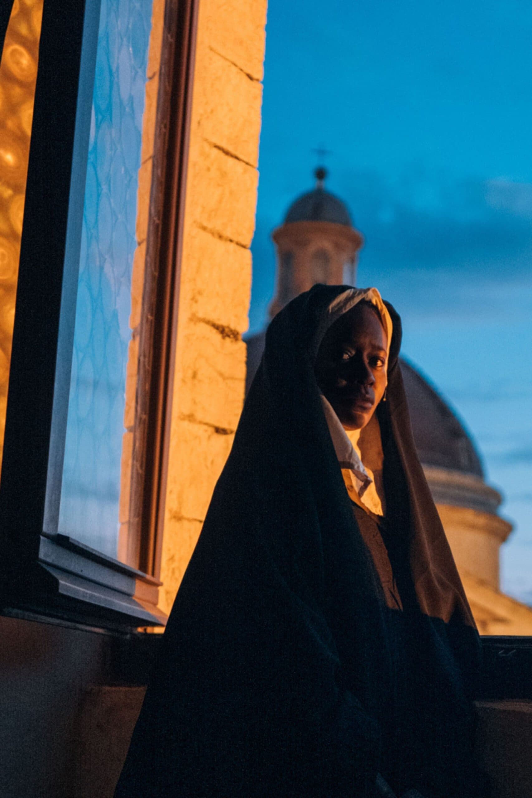 A film still from Run, Sister Joan shows a Black nun standing at an opened window. The side of her face is illuminated by the light from outside as she looks over her shoulder at the camera with a serious face.