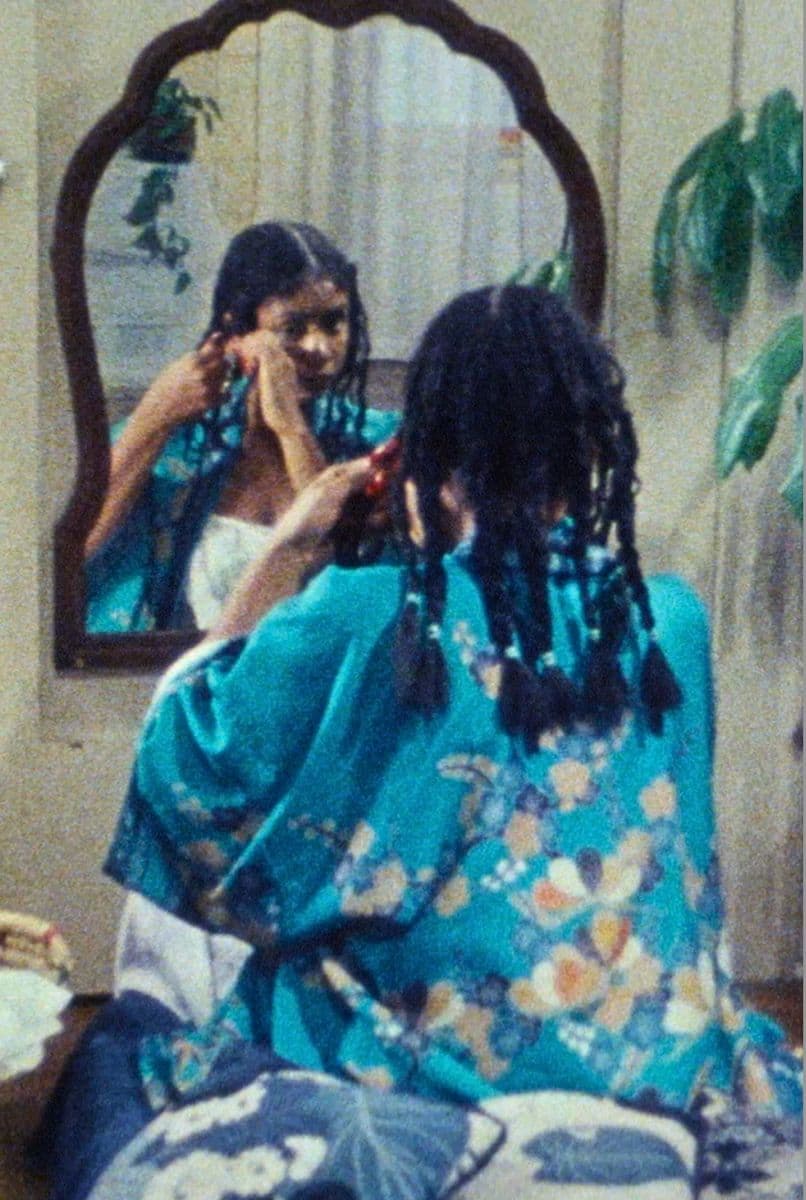 A film still from A Different Image shows a young Black woman sitting in front of a mirror, braiding her hair. She is wearing a blue outer layer with flower patterns and is sitting on a pile of white and blue pillows.