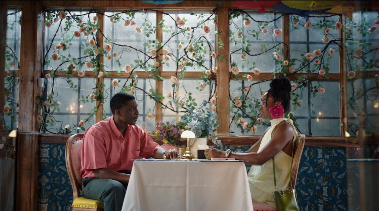 A film still from Call Of My Life shows a Black man and a Black woman sitting at a square table. The man is wearing a pink shirt and is looking toward the woman. The woman is looking back with a soft smile and is wearing a pale yellow dress and large pink flower earrings. There is a small lamp and two large bouquets of flowers on the table. Behind them, there is a wooden trellis wall with vines of pastel pink-orange roses.