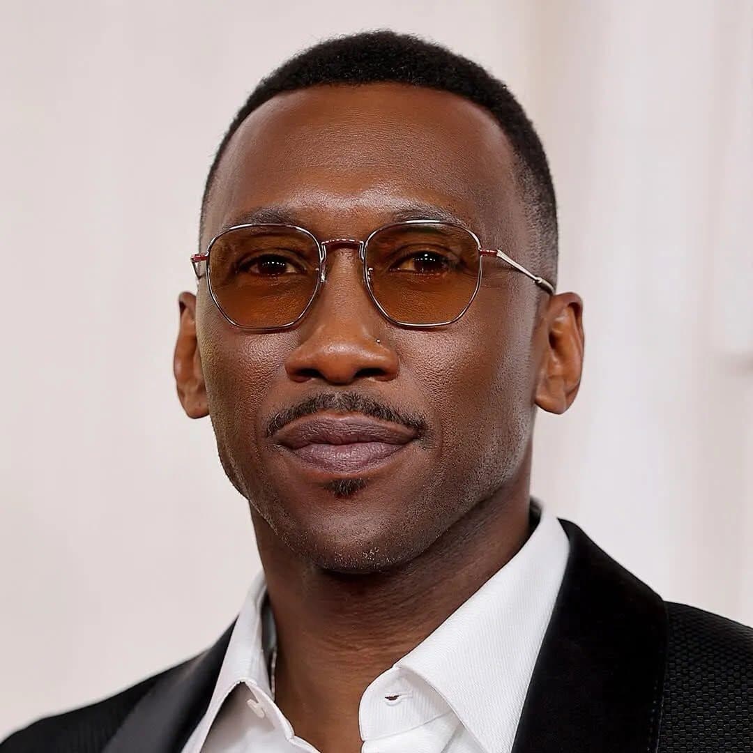 A headshot of Mahershala Ali. He is a Black man, he is wearing a suit, light-colored glasses, and has a soft smile.