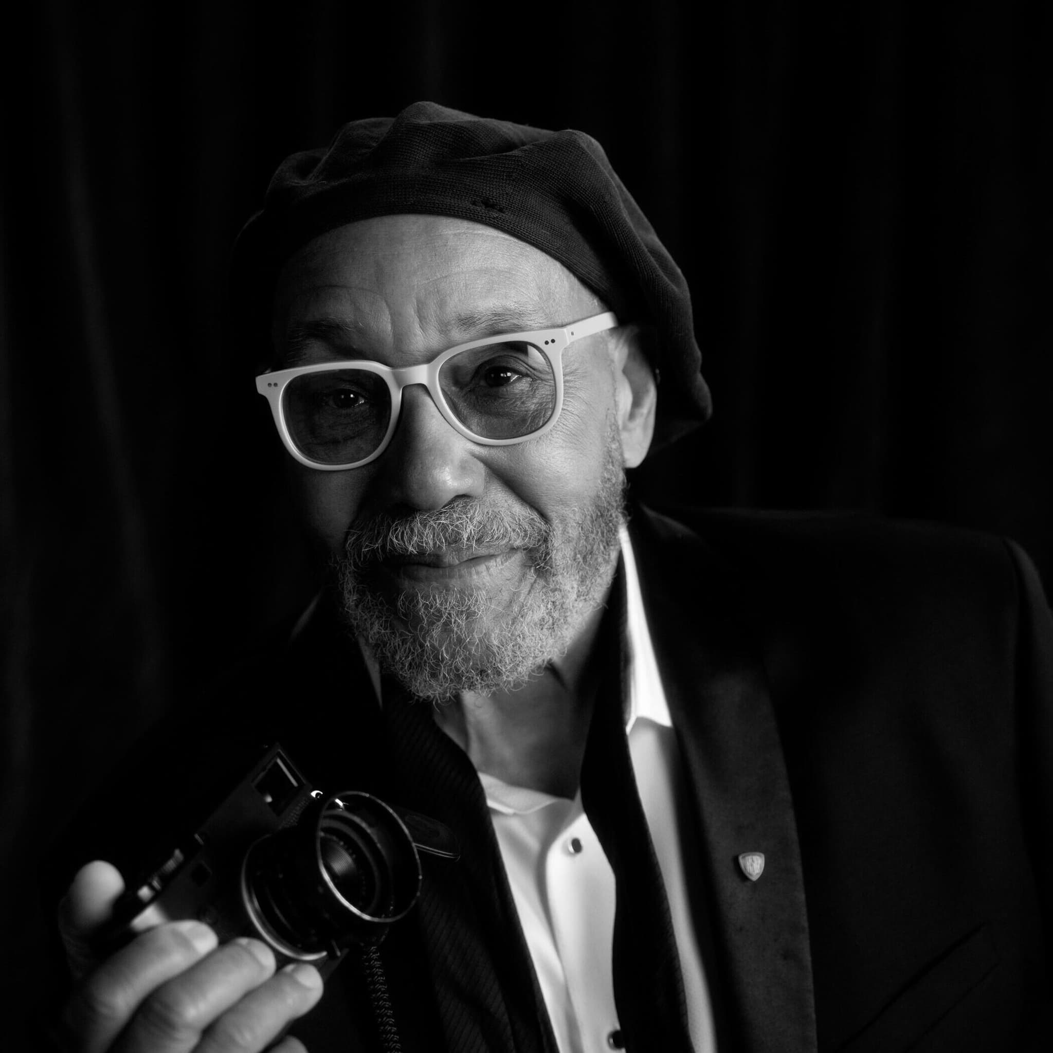 A headshot of John W. Simmons. He is a Black man. He has a soft smile and is wearing rimmed glasses and holding a camera.