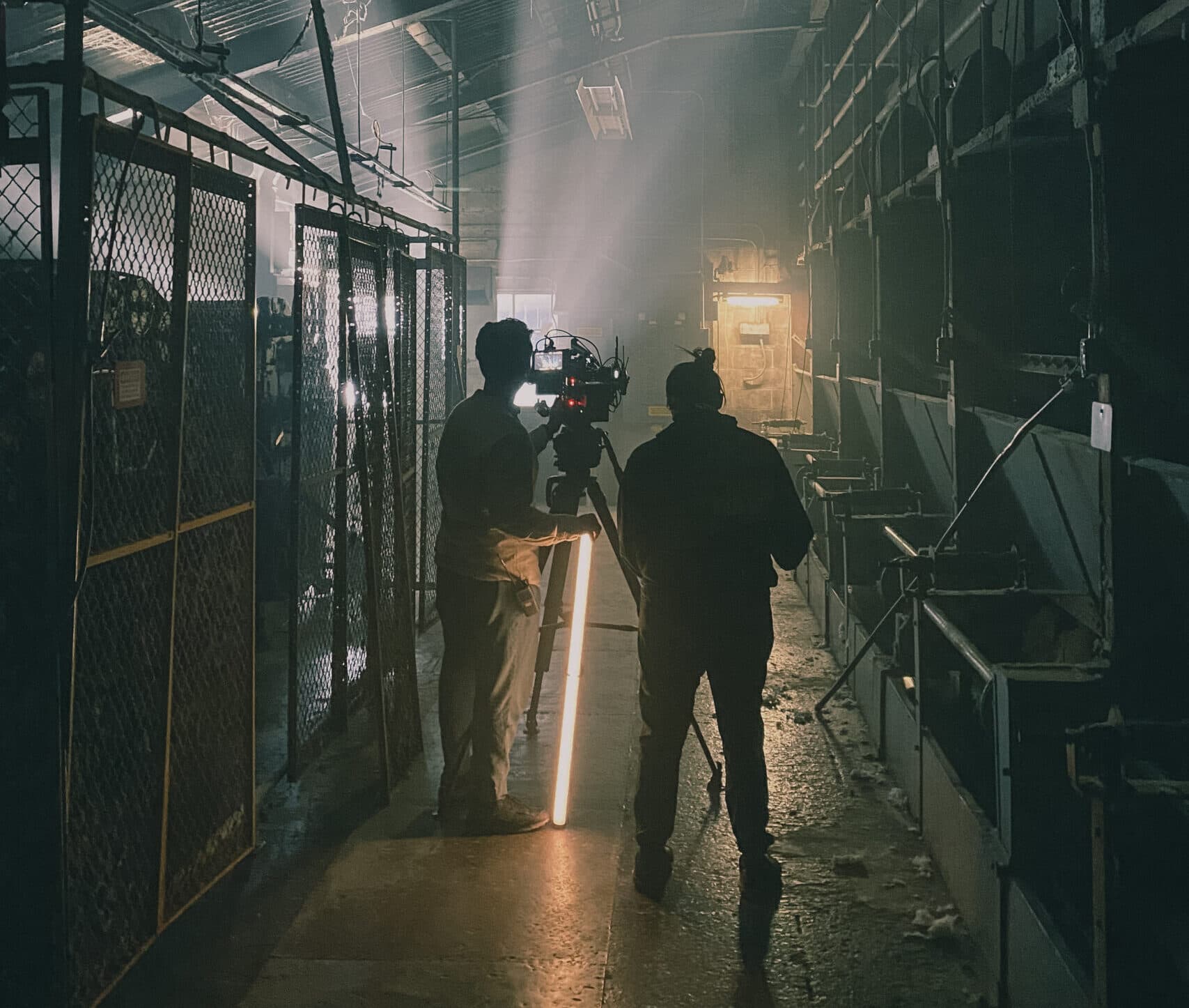 A photo of two people on set, they are in a dark warehouse standing behind a camera mounted on a tripod. They are backlit, there is a glow coming from an open door and a light in front of them.