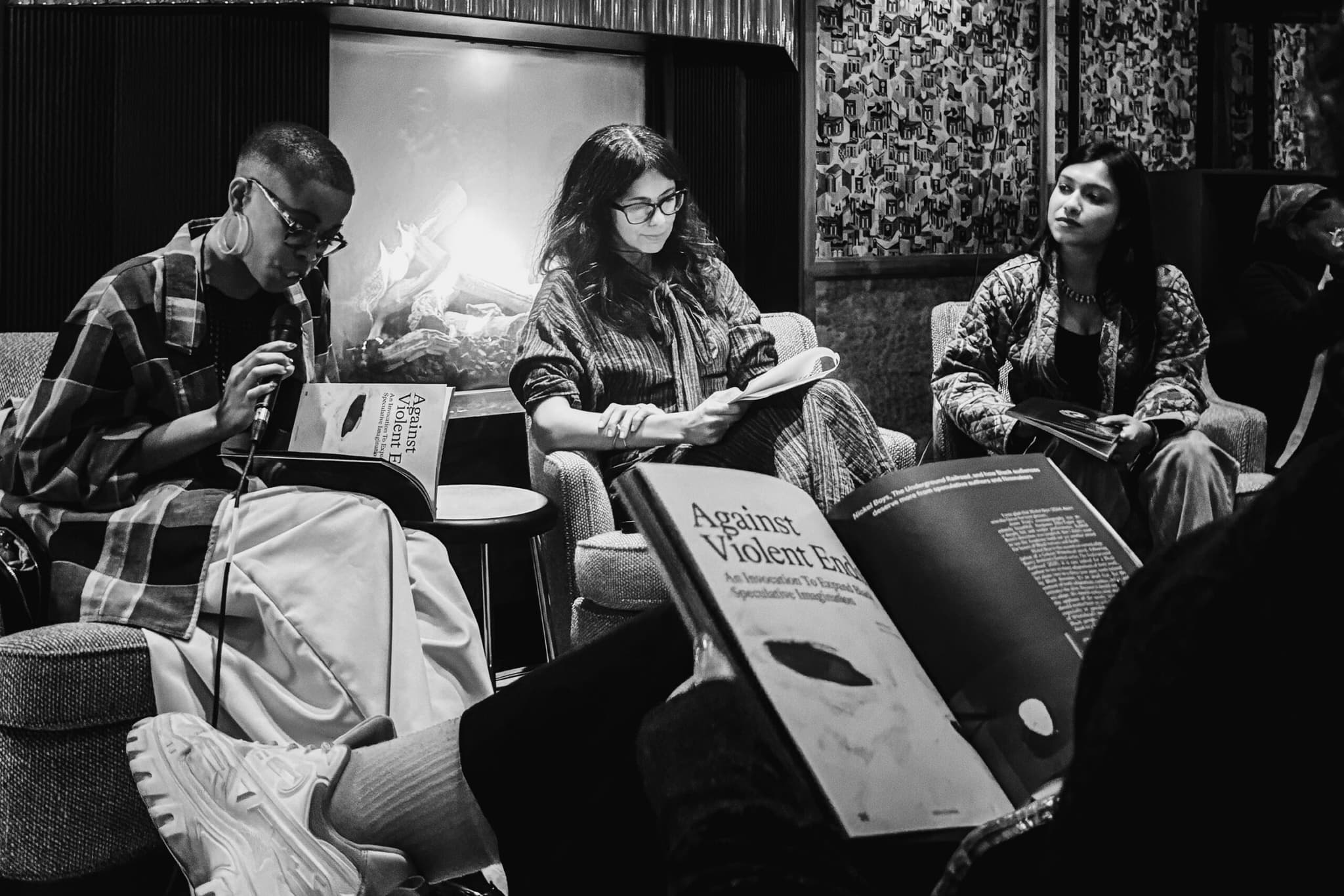 A black-and-white photo of people gathered for a reading. There are three people sitting in the front of the room, they have their journals open.