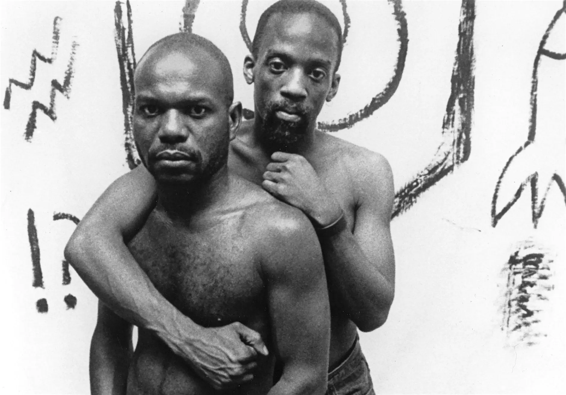 A still from Tongues Untied shows two Black men holding one another. They are not wearing shirts, and are looking at the camera with stern expressions.