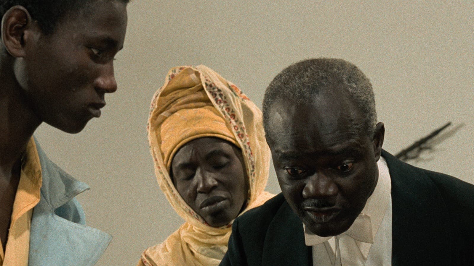 Film still of a young man in profile looking downward. Beside him, an older Black woman, her head wrapped in a tan head covering, has her eyes closed, as an older Black man stares at something below.