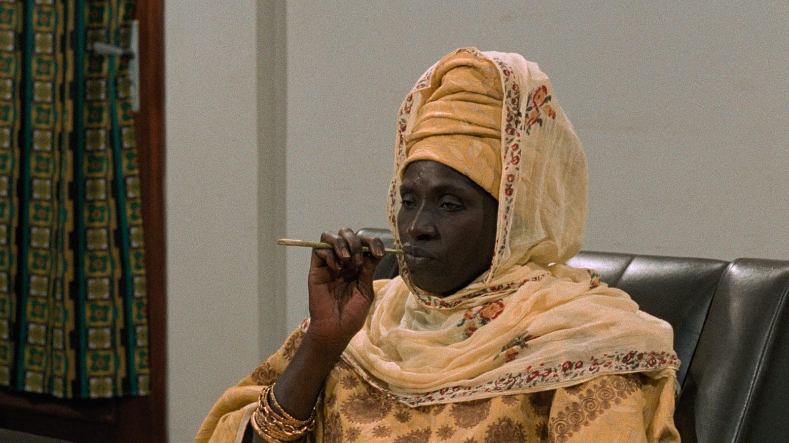 Film still of a Black woman seated in a dark leather chair, wearing a tan head wrap under a cream shawl embroidered with red flowers and a patterned dress. She holds a slim chewing stick to her lips, gold bangles stacked on her wrist, and gazes off to the side.