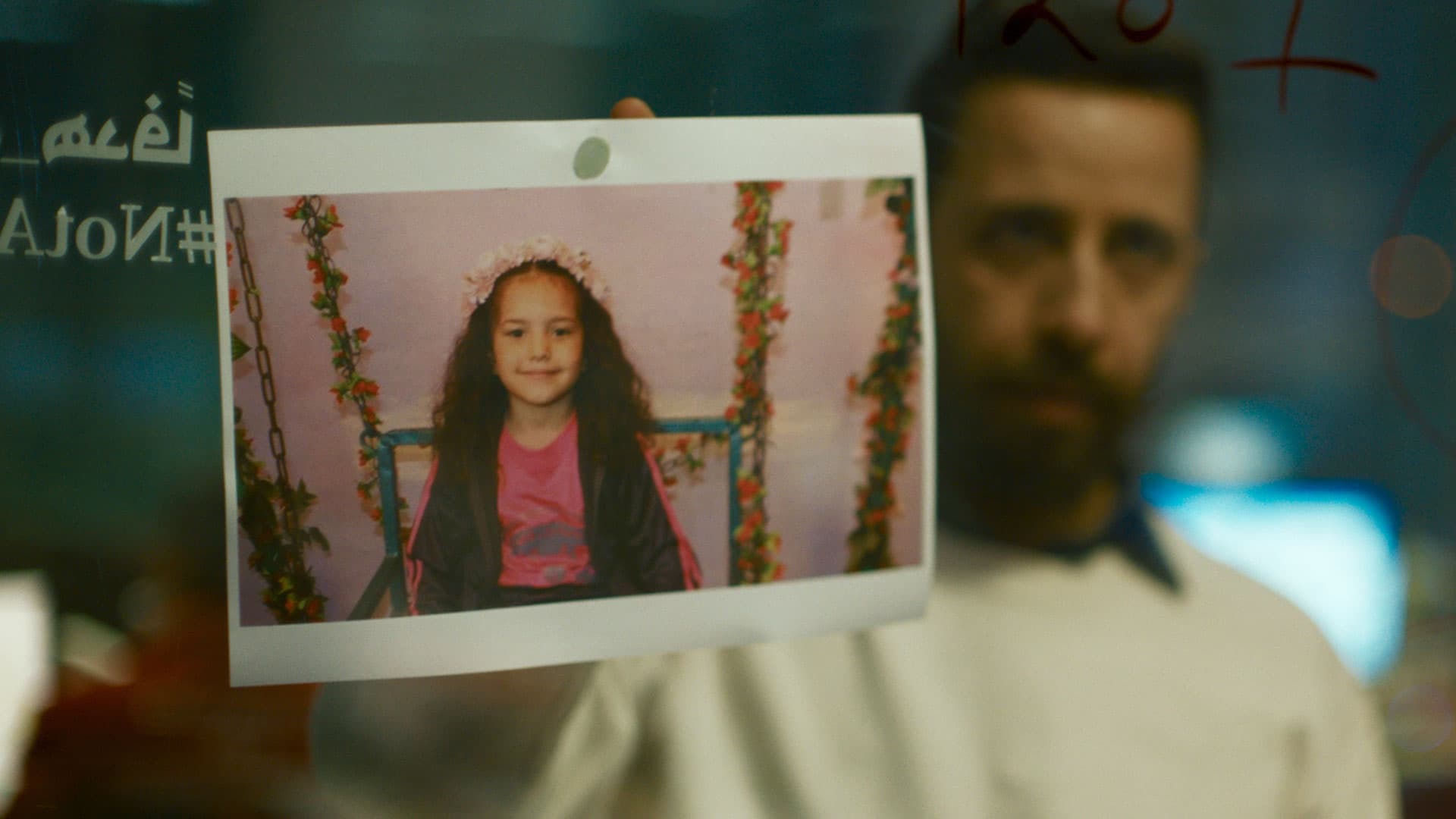 A man, who appears out of focus, holds up a photograph of a girl smiling with a flower crown on her head.