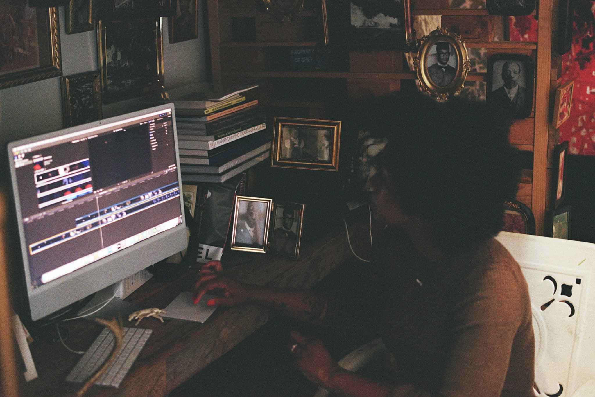 Color photograph of Allison Janae Hamilton at a wooden desk, holding a computer mouse and gazing into a monitor, surrounded by framed photographs and a stack of books