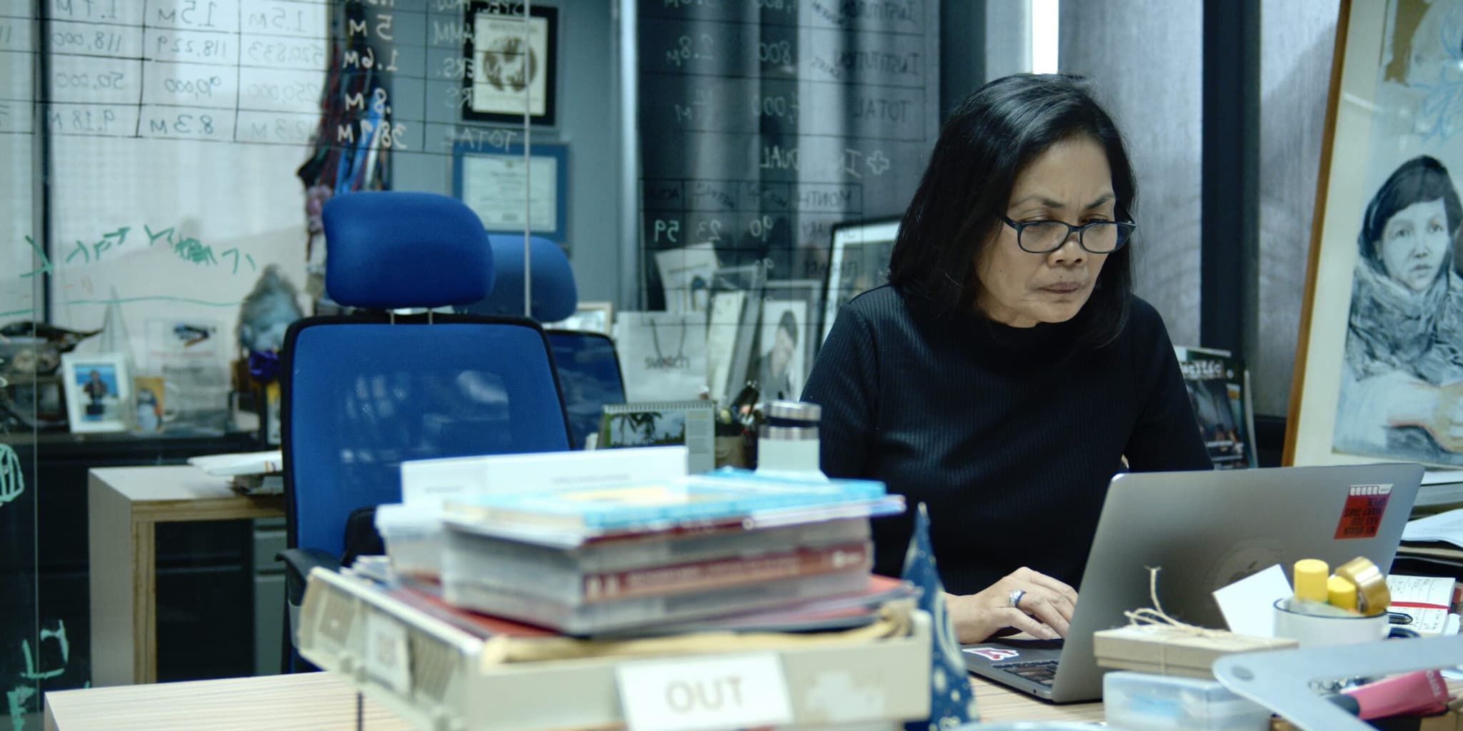 A Filipino woman wearing glasses types on a laptop at a desk piled high with books.