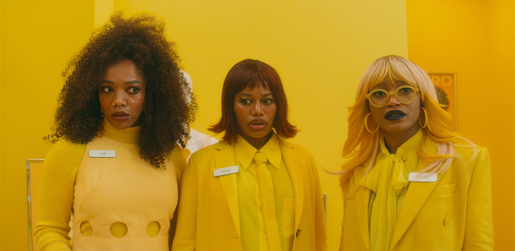 Film still of three Black women standing side by side in matching pale yellow coats. One has full dark curly hair, one a sleek dark bob, and one platinum hair with white-framed glasses.