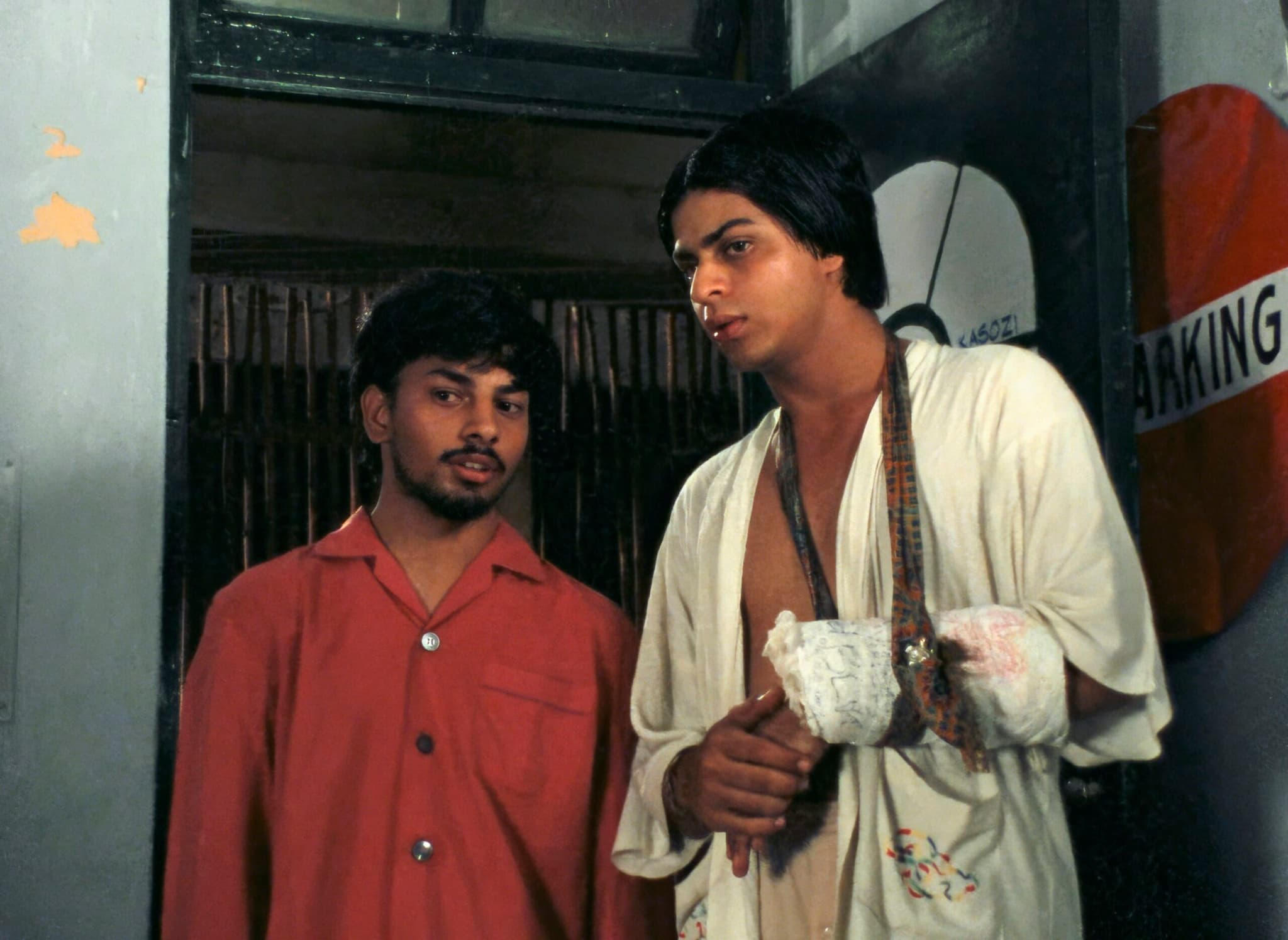 Two young men stand in a dim doorway; one in a red shirt, the other in an open cream robe with bandaged, ink-marked forearms.