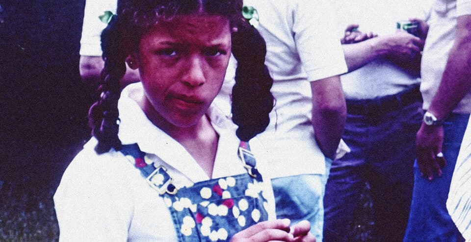 A photo of Michele Stephenson as a young girl. She is wearing denim overalls with polkadots over a white button down shirt. She has pig tails and a smirk on her face.