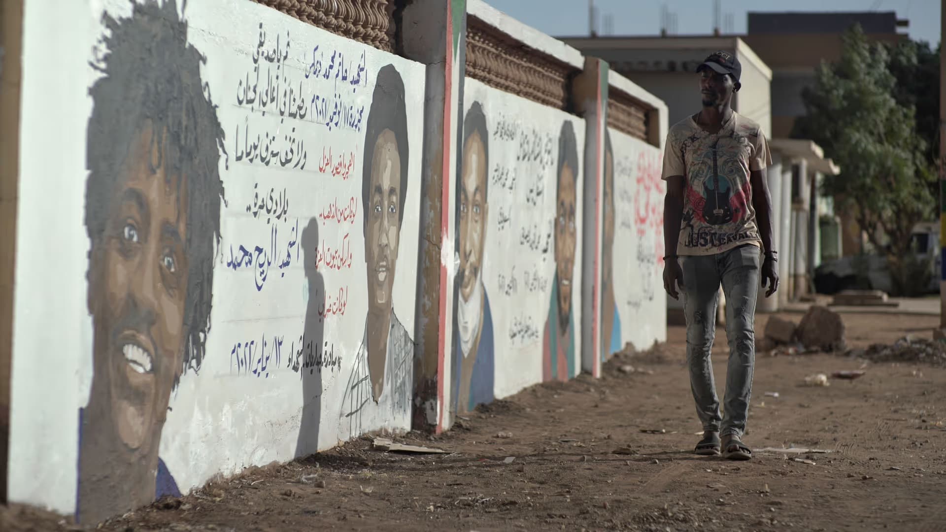 A man walks past a large white wall featuring mural portraits of young men alongside Arabic text.