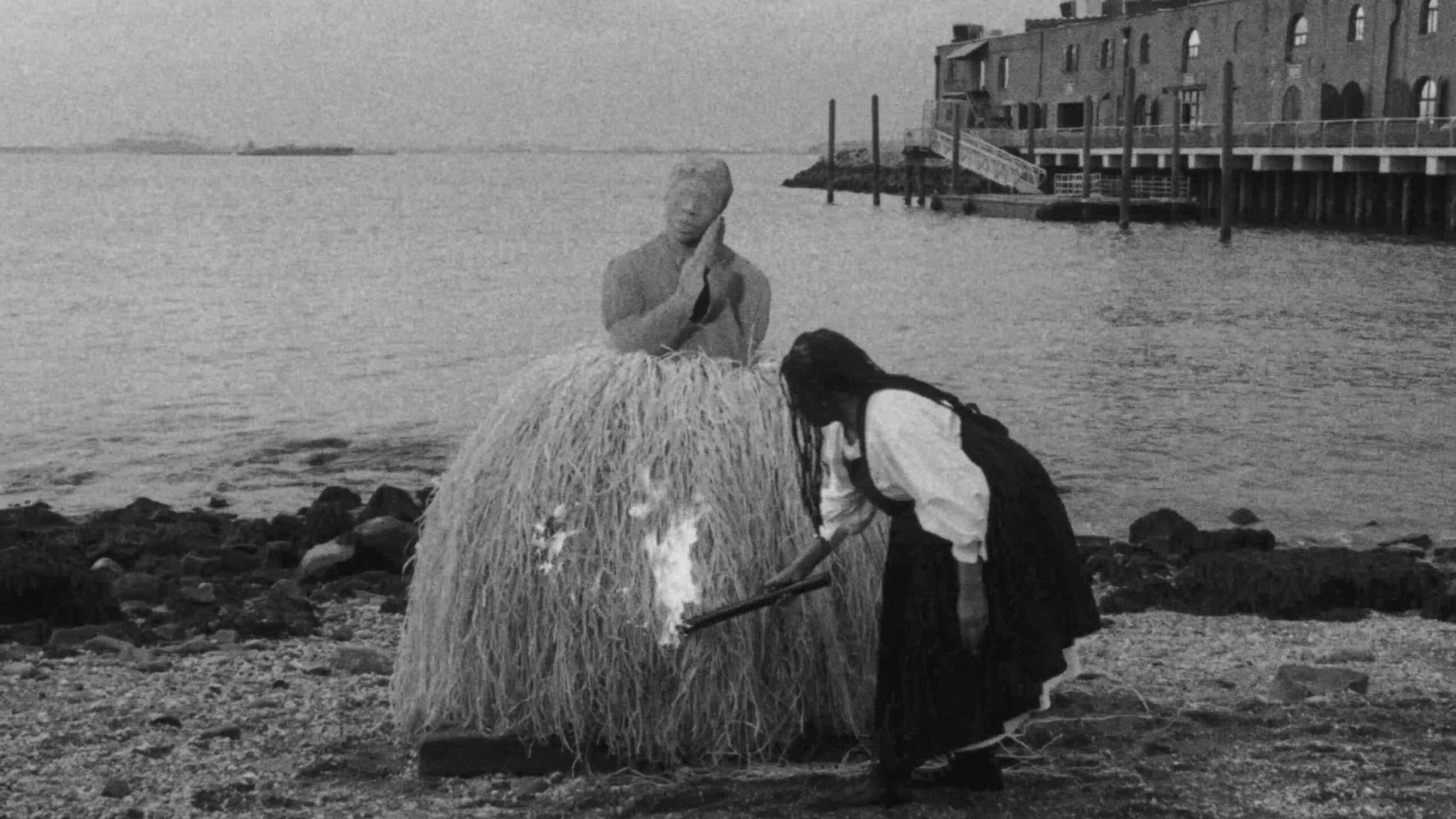 Black-and-white film still of the artist Simone Leigh in a black pinafore over a white blouse crouching low, holding a stick lit with fire to the base of one of her life-sized raffia-skirted sculptures.