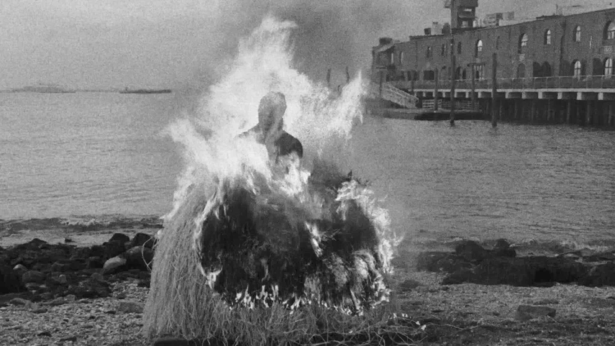Black-and-white film still of the artist Simone Leigh in a black pinafore over a white blouse crouching low, holding a stick lit with fire to the base of one of her life-sized raffia-skirted sculptures.