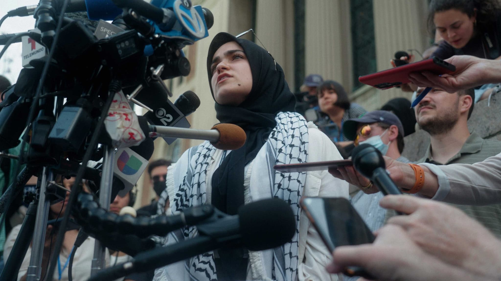 On Columbia University’s campus, a woman with a keffiyeh around her neck stands before a camera, while encircled by journalists holding microphones.