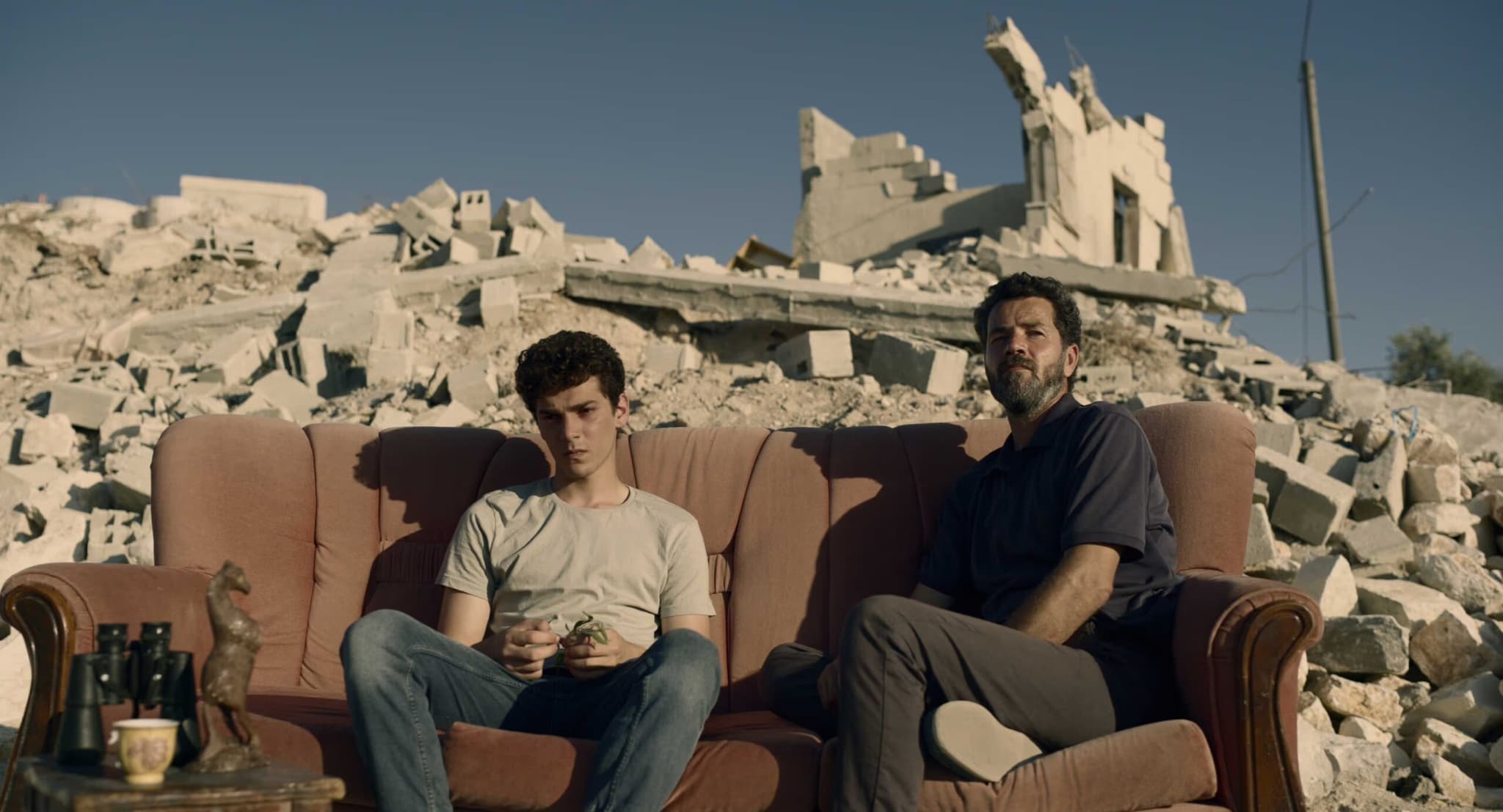 Two men sit on a dusty worn couch amidst rubble, in front of a house that appears destroyed by bombing.