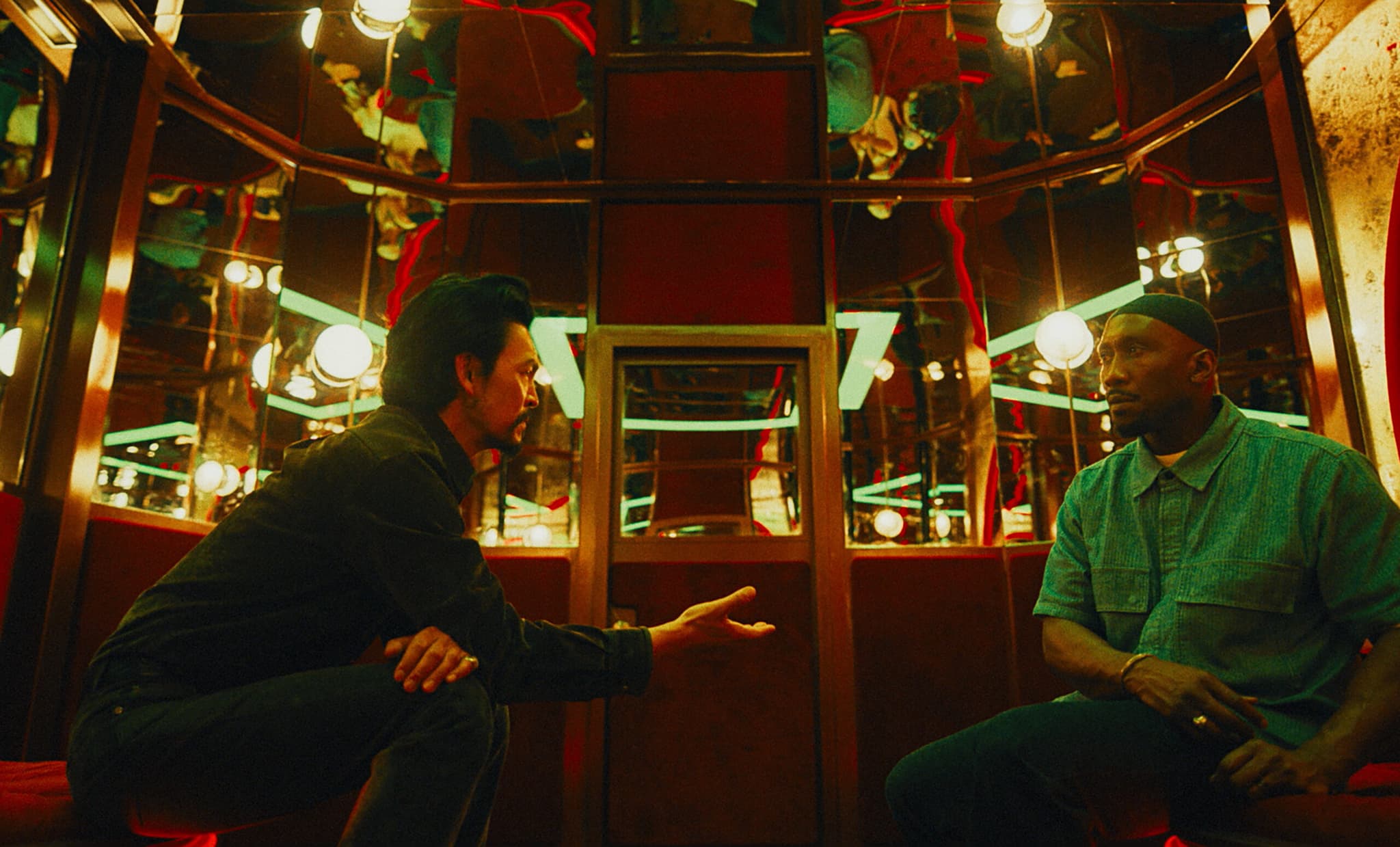 A still from Your Mother, Your Mother, Your Mother. It shows two men in conversation, they are in dimly lit room with mirrors and glass bulb lights. They are sitting on red velvet benches.