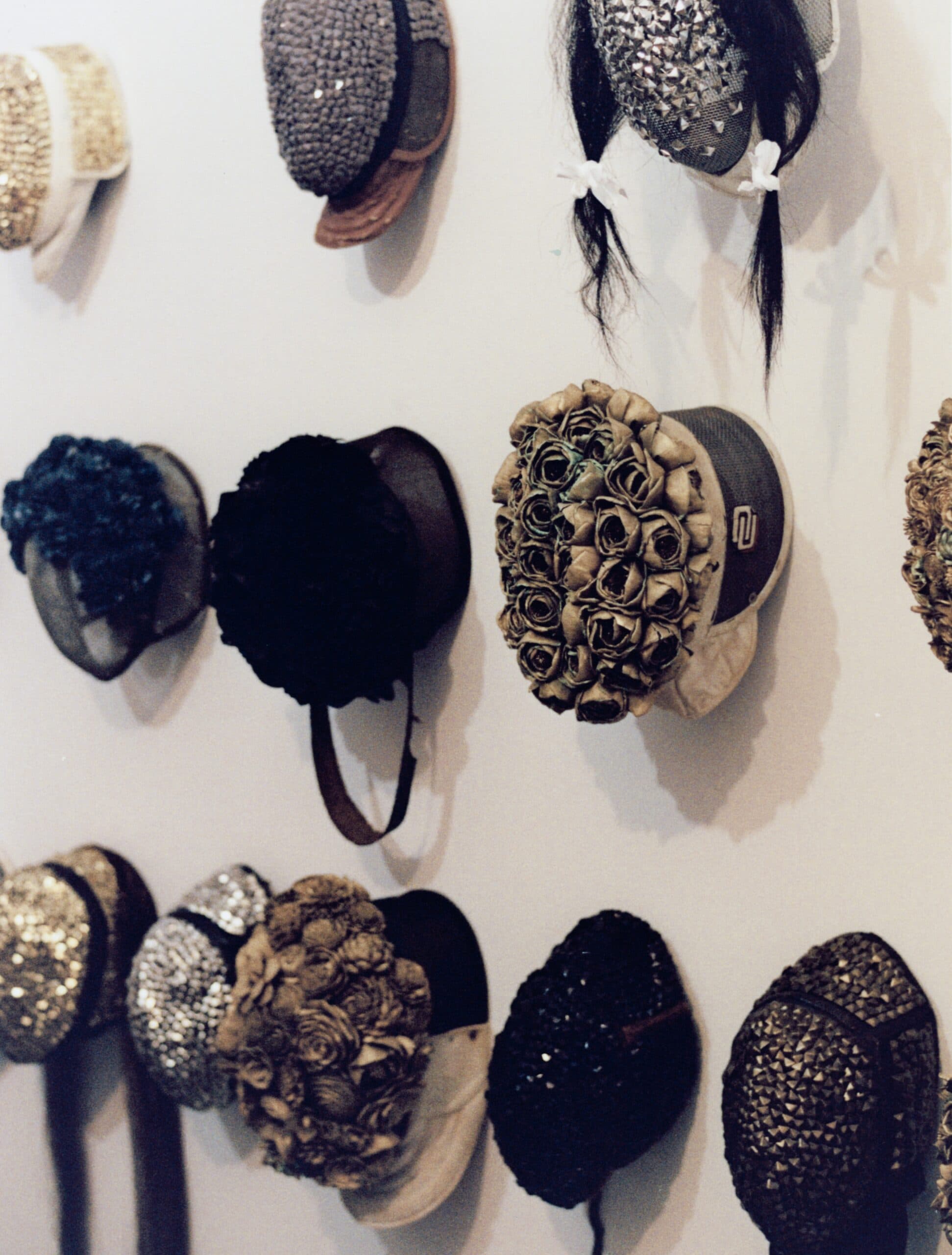 Vintage hats, some sequined, some designed with gold roses, sequins and studs hang on a white wall in three rows.